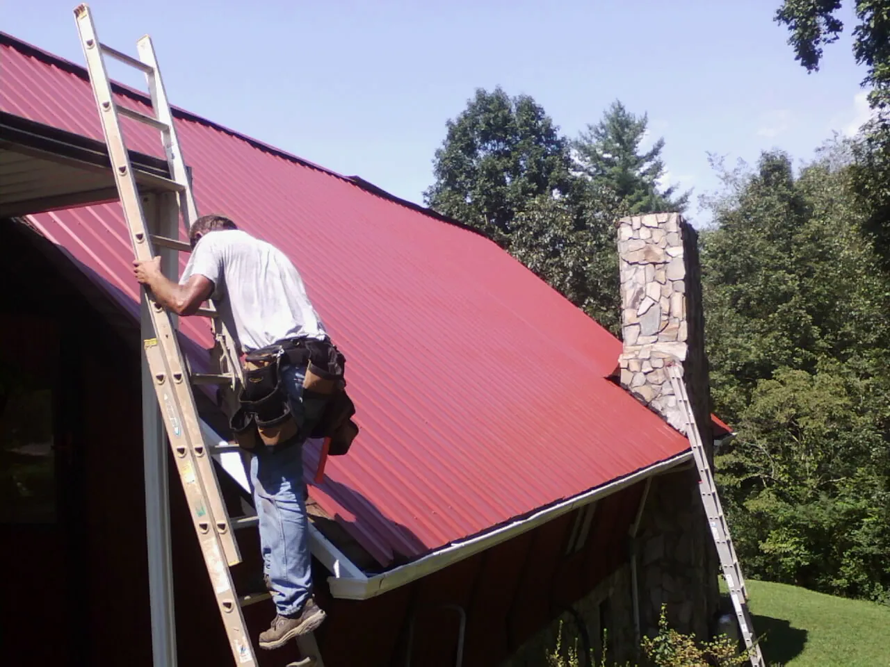 Roofer on a ladder beside a red metal roof and stone chimney — Soffit & Fascia Repair by Roofing Brothers in Lattimore